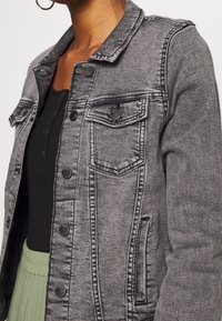 Grå denimjacka med knäppning fram, två fickor med lappar och kontrasterande sömnadsdetaljer. Har en klassisk krage och en strukturerad passform.