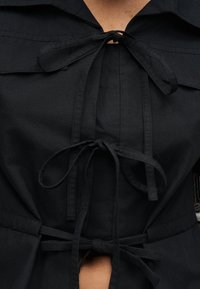 Κοντινό πλάνο μαύρου ενδύματος με τρεις υφασμάτινους δεσμούς κουμπωμένους μπροστά πάνω σε γυμνό δέρμα, που δείχνει γιακά και μερική μέση.