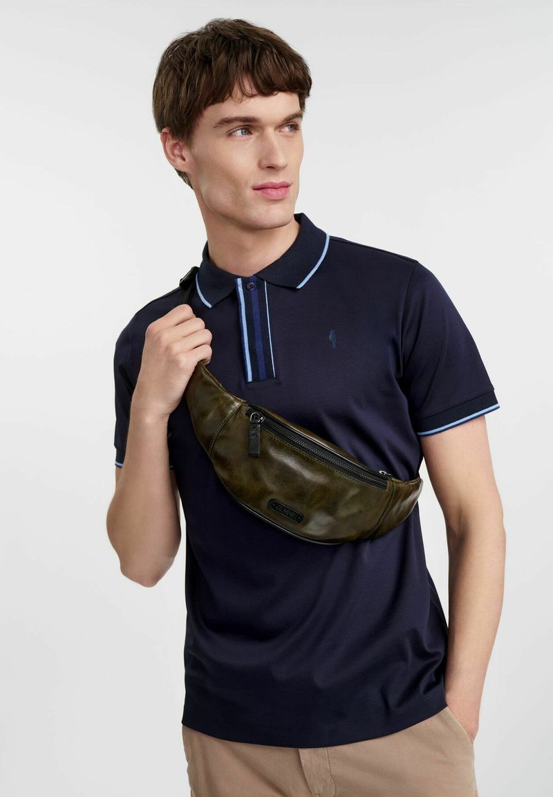 Zielona skórzana torba na ramię o zaokrąglonym kształcie i kieszeni na zamek, noszona na granatowej koszulce polo z jasnoniebieskimi akcentami i subtelnym logo.