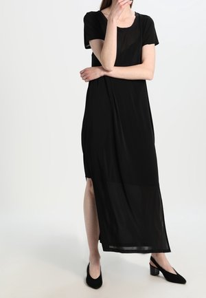 Kvinde iført en lang sort kjole med korte ærmer og sideskæring, stående med den ene hånd på hagen og armene krydsede, sorte sko med lav hæl.