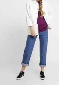 Chaqueta de mezclilla blanca, camiseta burdeos con gráfico amarillo, jeans azules con dobladillo enrollado, zapatillas negras de caña alta con suela blanca.