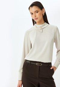 Blusa beige clara de manga larga con cuello y un detalle de botón, combinada con pantalones marrones oscuros y un cinturón delgado con hebilla dorada.