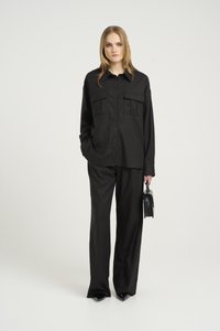 Camicia nera con bottoni, dotata di due tasche sul petto, abbinata a pantaloni neri a gamba larga. La modella tiene una piccola borsa nera. Tessuto liscio.