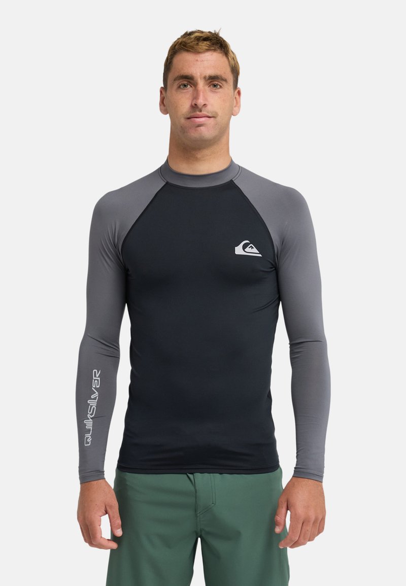 Rash guard à manches longues noir et gris avec une coupe ajustée. Présente un logo sur la poitrine et la manche. Tissu doux et extensible, adapté aux sports nautiques.