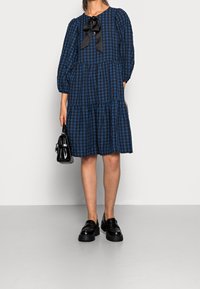Blau- und schwarz kariertes Kleid mit rundem Ausschnitt, Puffärmeln und einer Schleifendetail, ergänzt durch eine schwarze Handtasche und klobige schwarze Schuhe.