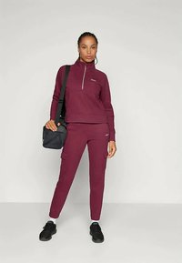 Woman standing wearing a matching burgundy zip-up sweatshirt and cargo joggers, black sneakers, and carrying a black shoulder bag.