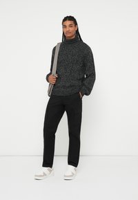 Chunky strikket sort sweater med høj krave, parret med sorte cargo bukser. Hvide sneakers med grå accenter. Modellen holder en grå skuldertaske.