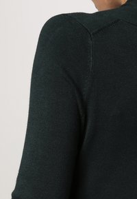 Mørkegrønn strikket sweater med en glat tekstur, der har en tætsiddende krave og indsyede ærmer, som fremviser ribbede detaljer ved sømmene.