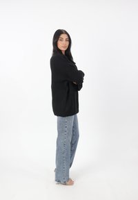 Maglione nero oversize con maniche lunghe, abbinato a jeans a zampa d'elefante blu chiaro e sandali con tacco trasparente, in piedi su uno sfondo bianco.