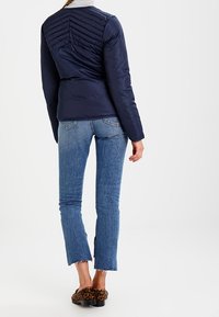 Marine pufferjack met geborduurd detail op de rug, gedragen over lichtblauwe jeans met rafelige zoom en slip-on schoenen met luipaardprint.