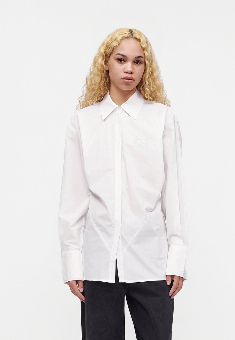 Jeune personne aux longs cheveux blonds bouclés, portant une chemise blanche boutonnée et un pantalon noir, debout devant un fond blanc uni.