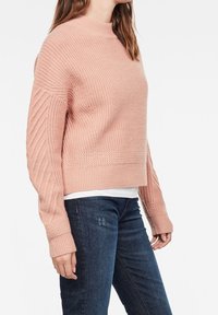 Ljust rosa stickad tröja med hög hals och ribbad textur, med ribbade ärmar och en kort passform. Kombineras med mörka jeans.