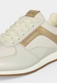 Baskets Michael Kors blanches et beiges avec lacets, logo sur la bande latérale et sur la languette, semelle texturée visible sur le bord inférieur.