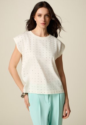 Donna con top bianco senza maniche con piccoli puntini neri e pantaloni a vita alta azzurri, in posa davanti a uno sfondo beige.