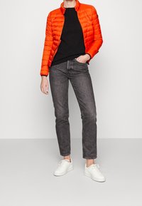 Veste orange rembourrée avec fermeture éclair, pull noir, jeans gris coupe droite et baskets blanches. Tissu lisse et léger avec un design matelassé.