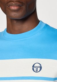Lühikeste varrukatega sinine puuvillane T-särk valgete ja helesiniste triipudega, mille rinnal on tumesinises broderingus ringikujuline logo.