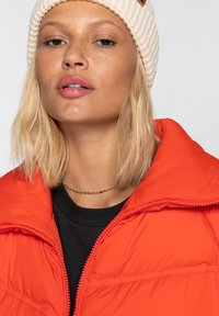Giacca a vento arancione brillante con colletto alto e zip, abbinata a una maglietta nera e un berretto lavorato a maglia color crema, con un piccolo dettaglio del logo.