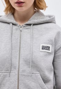 Κοντινό πλάνο ενός ατόμου που φοράει ανοιχτό γκρι φούτερ με φερμουάρ και κορδόνια, με ένα ορθογώνιο ταμπελάκι λογότυπου "Nørgaard" στο στήθος.