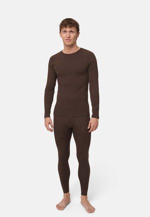 Homme debout pieds nus portant un haut à manches longues marron foncé et des leggings moulants assortis sur fond blanc uni.