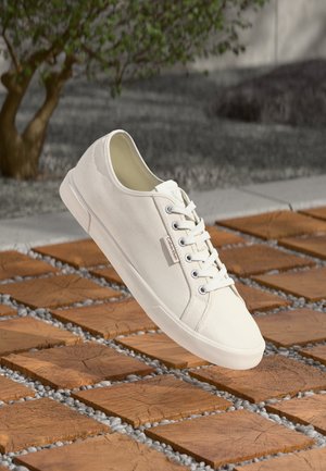 Baskets blanches en toile avec lacets flottant au-dessus d'un sol extérieur en carreaux de bois et galets, petite étiquette Calvin Klein sur le côté.