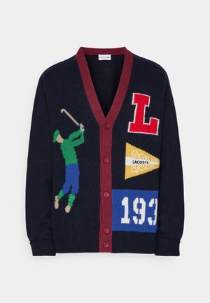 Tmavý kardigan s bordó lemováním knoflíků, s motivem golfisty, červeným písmenem „L“, žlutou vlaječkou Lacoste a bílým „1933“ na modré nášivce.