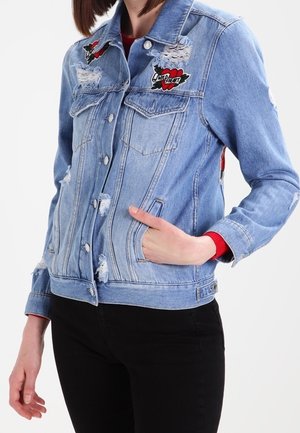 Giacca in denim blu chiaro con dettagli distressed, due tasche sul petto e toppe ricamate. Presenta una chiusura a bottoni e un design aderente.
