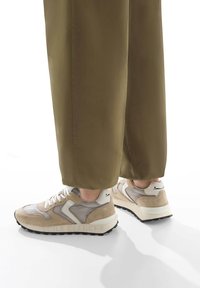 Sneaker beige e grigio realizzata in materiali scamosciati e mesh, con una suola nera chunky e dettagli bianchi. Abbinata a pantaloni affusolati verdi oliva.
