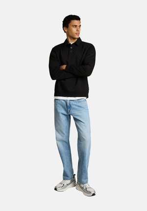 Männliches Model steht mit verschränkten Armen, trägt ein schwarzes langärmeliges Polo, hellblaue Jeans und weiße Sneakers vor weißem, einfarbigem Hintergrund.