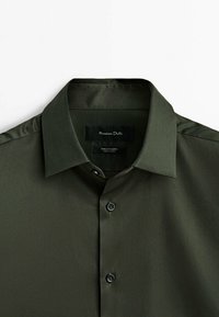 Camicia verde scuro con bottoni, realizzata in tessuto pregiato. Presenta un colletto classico, bottoni neri e una trama sottile. L'etichetta mostra il nome del marchio.