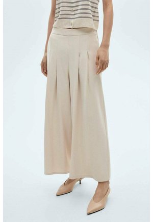 Frau in beigen weiten, gefalteten Hosen, nackten, spitz zulaufenden Kitten-Heels und einem ärmellosen, gestreiften Oberteil mit Knopfleiste.