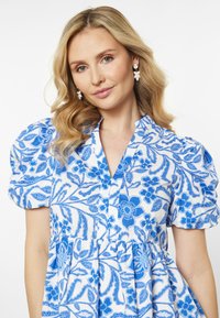 Rochie albă cu modele florale și paisley în nuanțe de albastru, mâneci scurte tip bufantă, decolteu cu deschidere în față și detalii pliate la talie.