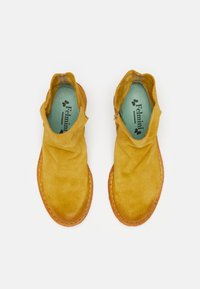 Botas de tobillo de ante amarillo con una parte superior suave y drapeada, suela de goma naranja y plantilla azul con el logotipo de la marca.