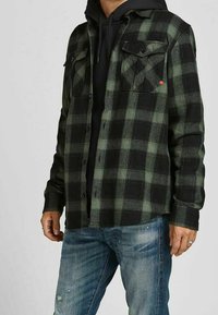Camicia flanella a quadri verdi e neri con due tasche sul petto, chiusura con bottoni e maniche lunghe indossata sopra un hoodie nero.