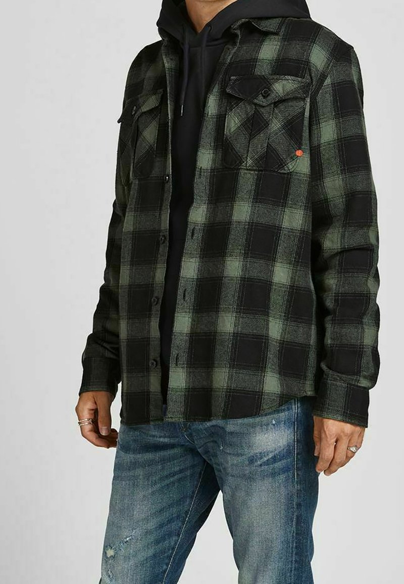 Camicia flanella a quadri verdi e neri con due tasche sul petto, chiusura con bottoni e maniche lunghe indossata sopra un hoodie nero.