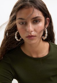 Silberne Creolen mit einem strukturierten, abstrakten Design. Das Model trägt ein dunkelgrünes Oberteil. Lange, wellige Haare verdecken teilweise das Gesicht.