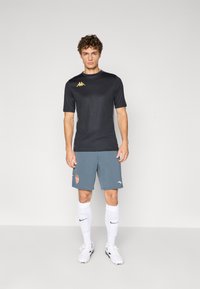Zwart sport T-shirt met korte mouwen, grijze sportshorts met een logo, en witte sokken. Model draagt voetbalschoenen. Eenvoudig ontwerp, casual pasvorm.