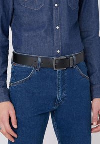 Sorte læderbælte med en rektangulær spænde, båret over en denimskjorte, parret med blå jeans. Har bæltetikker og syede detaljer.