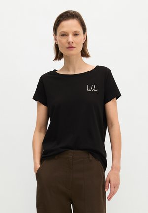 Femme aux cheveux bruns courts portant un t-shirt noir avec l’inscription « hello » sur la poitrine et un pantalon marron, debout devant un fond blanc uni.