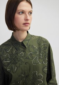 Olijfgroene button-up shirt met bloemenborduursel en metallic accenten. Heeft een puntige kraag en een ontspannen pasvorm. Getextureerde stof.