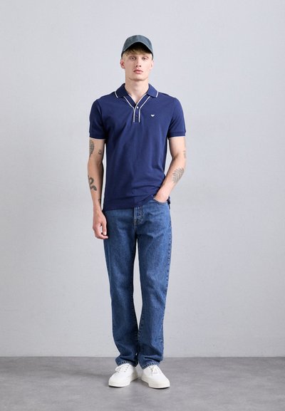 Polo de marină cu dungi albe, combinat cu blugi bleu deschis și adidași albi. Modelul poartă, de asemenea, o șapcă albastră.