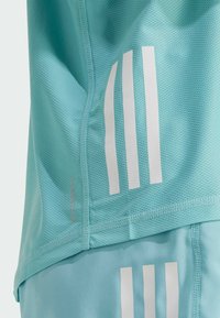 Shorts atletici de culoare albastru deschis, cu o textură din plasă, având accente albe cu trei dungi și branding "AEROREADY" pe lateral.