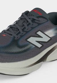 Σκούρο γκρι και μαύρο παπούτσι τρεξίματος New Balance με λευκή ενδιάμεση σόλα, γκρι κορδόνια και κόκκινο λογότυπο "ellipse" στη γλώσσα.