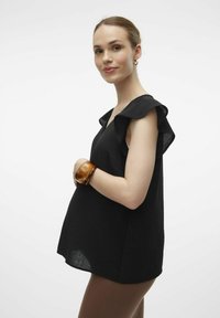Sort ærmeløs top med rynkede skuldre, struktureret stof og en løs pasform. Båret med et brunt armbånd og brune leggings.