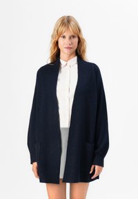 Gilet en mailles bleu marine à coupe décontractée, avec une ouverture sur le devant et deux poches avant. Il présente une texture côtelée et des manches longues.