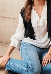 Blusa bianca con dettagli in pizzo e pois, abbinata a jeans in denim blu con borchie argentate lungo i lati, indossata con scarpe marroni.