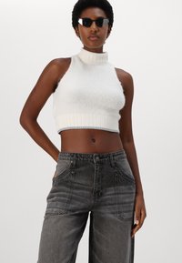 Balts adīts īsais tops ar augstu apkakli un ribotu apakšmalu, kopā ar pelēkiem platajiem džinsiem, kuriem ir priekšējās kabatas un izbalējusi tekstūra.