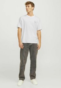 Hvid kortærmet t-shirt med rund hals og et kraniumgrafik på venstre bryst, parret med mørke denimjeans og hvide sneakers.