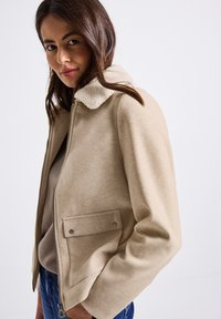 Žena s dlouhými hnědými vlasy, oblečená v béžové bundě s kožešinovým límcem, béžovém topu a modrých džínách, stojící na bílém pozadí.