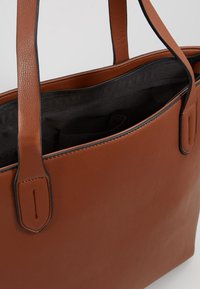 Hnědá kožená taška přes rameno s texturovaným povrchem, černým interiérem, robustními madly a kontrastním černým lemováním podél horního okraje a kapes.