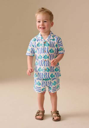 Naeratav väikelaps, kes kannab sinakasroheliste kalade ja lainete mustriga sobivat lühikeste varrukatega särki ja lühikesi pükse, seistes beežil põrandal.